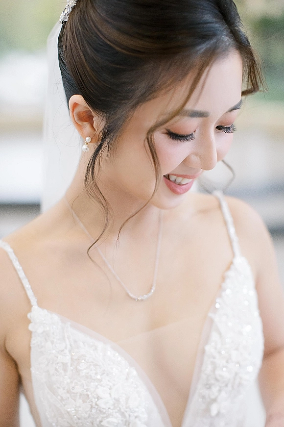 Bridal portrait of a bride looking down in soft window light, wearing a lace wedding dress and veil against blurred greenery backdrop