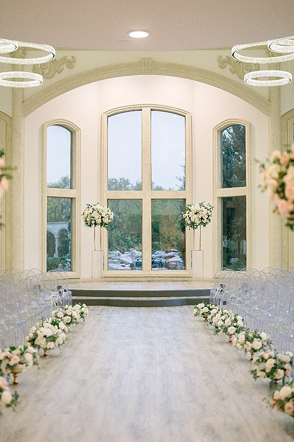 Ceremony setup for an indoor wedding ceremony with wedding aisle flowers, clear acrylic chairs, and ring chandeliers before arched windows