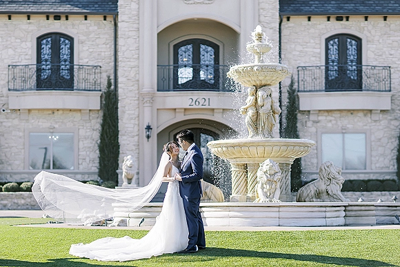 Couple portrait of bride and groom holding hands by a fountain, her cathedral veil blowing behind a strapless dress on a stone mansion lawn