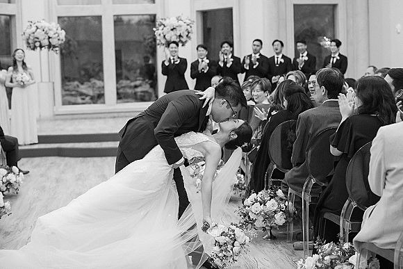 Wedding kiss portrait with a ceremony kiss dip as the bride in veil holds her bouquet while guests applaud in an indoor space with large windows