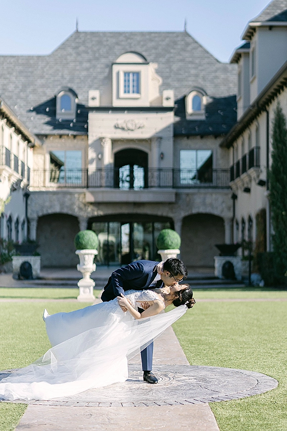 Wedding kiss portrait of groom dipping bride in lace dress and cathedral veil on a stone walkway before a grand estate facade in daylight