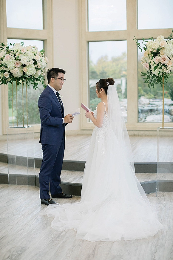 Wedding vows as bride and groom read from personal vow books, her cathedral veil glowing in natural light by large windows and florals
