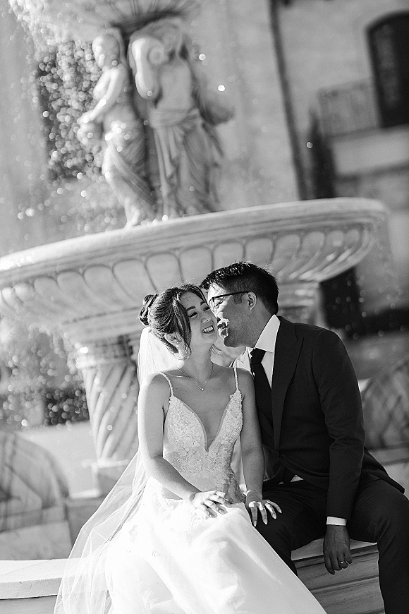 Couple portrait in a black and white wedding photo, bride in lace dress and veil laughing with groom in glasses by a stone fountain