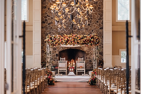 Ceremony setup with a floral arch framing a stone fireplace, white aisle runner lined with arrangements, candles, chandelier and string lights indoors