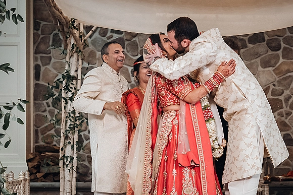 Wedding kiss at a hindu ceremony as bride in red lehenga and veil shares a kiss with groom in embroidered sherwani under canopy draping