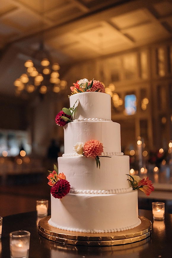 Wedding cake with smooth white buttercream, four tier wedding cake topped with dahlias and greenery on a candlelit stand in a chandelier-lit hall