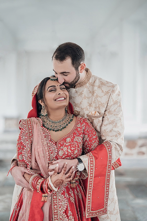 Couple portrait sharing a forehead kiss pose, bride in red lehenga and groom in cream sherwani against a simple white wall indoors