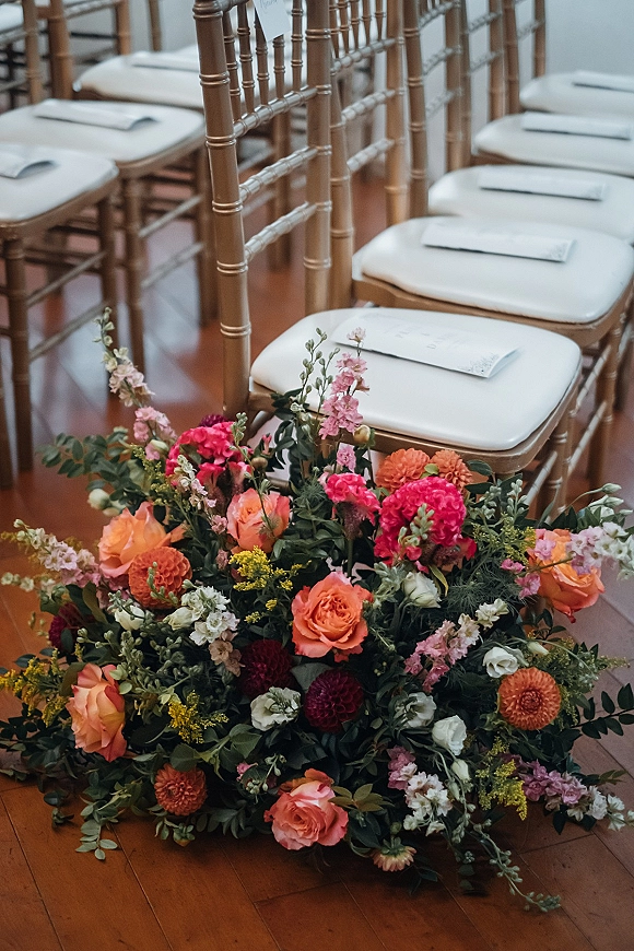 Ceremony aisle flowers in a lush ground arrangement with roses, dahlias, and greenery beside gold chiavari chairs on a wood floor indoors