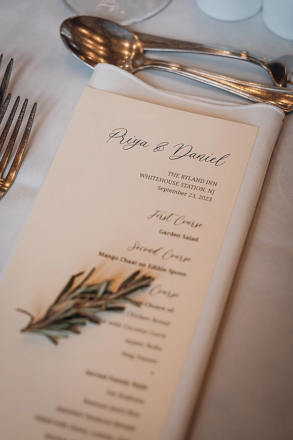 Wedding menu card with reception menu card in calligraphy on a white napkin beside silver flatware and a greenery sprig on white tablecloth