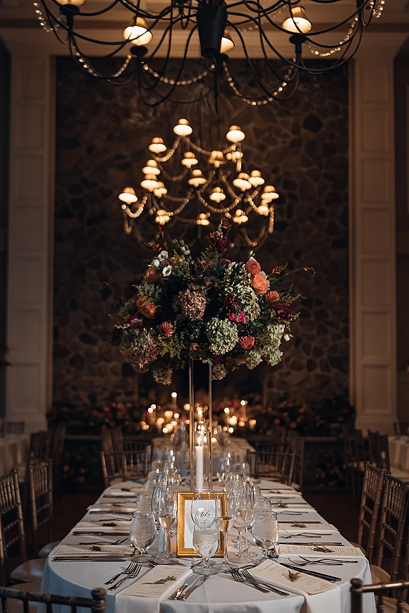 Reception tablescape with long head table decor, tall floral centerpiece in clear glass vase, candles, chandeliers, and string lights by a stone fireplace