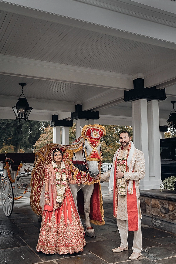 Wedding couple portrait of an Indian wedding couple in red lehenga and sherwani with flower garlands beside a decorated horse under a lantern-lit portico