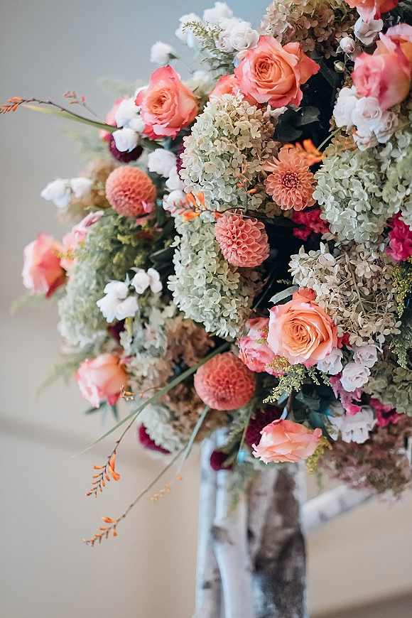 Wedding floral arrangement with peach rose wedding flowers, hydrangea, dahlias, and greenery on a stand against a neutral indoor wall