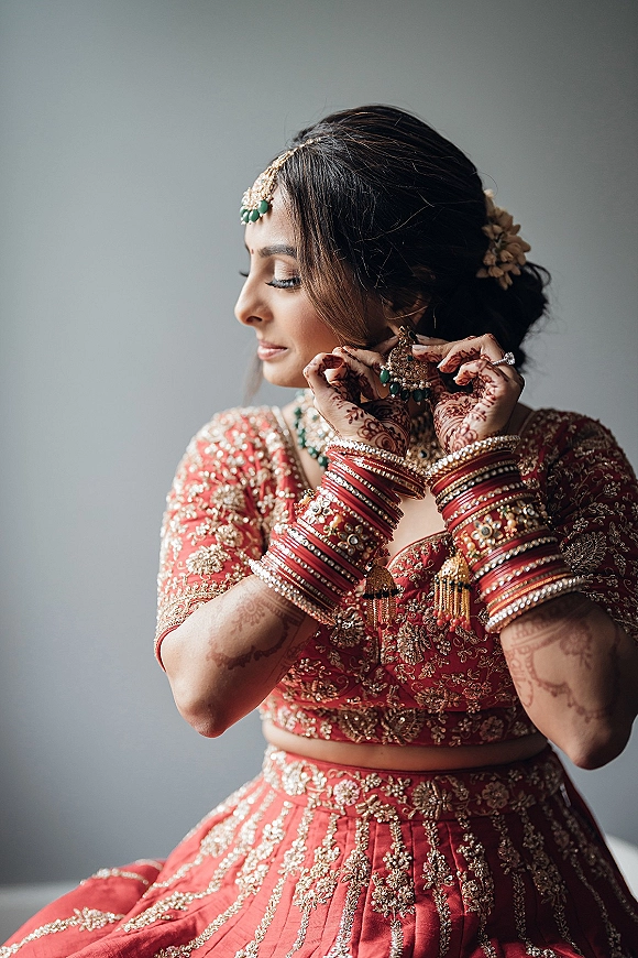 Bridal portrait of a South Asian bride in a red bridal lehenga, adjusting chandelier earrings with mehndi hands against a plain gray wall