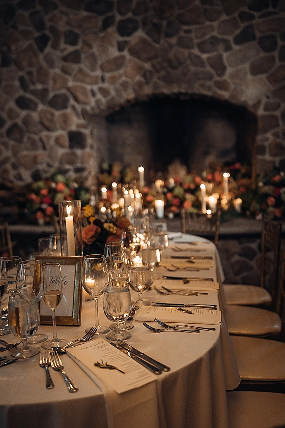 Reception tablescape with taper candles and votives along white linens, floral centerpieces, and place cards set before a stone fireplace