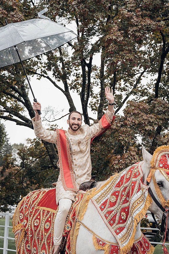Baraat groom on a white horse in an embroidered sherwani and red stole, holding a clear umbrella, with trees and fence behind.