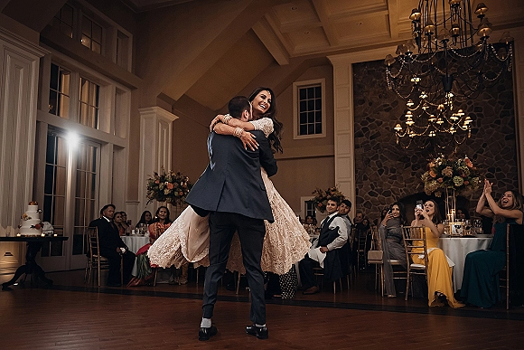 First dance as groom lifts bride in her wedding gown on a wood dance floor beneath a chandelier, guests cheering in a ballroom