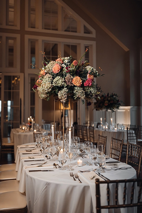 Reception tablescape with a wedding table centerpiece of tall roses and greenery, taper candles, and place cards in a bright ballroom