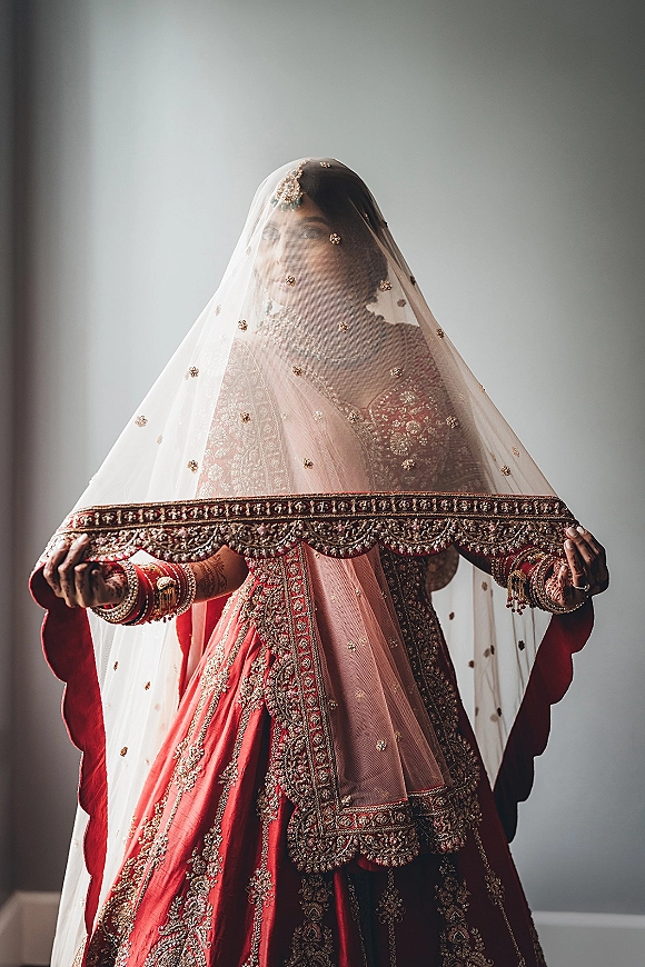 Bridal portrait of a bride in a red lehenga with dupatta over face, gold jewelry and henna hands, lit by window light indoors