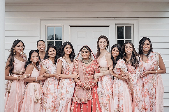 Bridesmaids portrait with Indian bride with bridesmaids, bride in red lehenga and gold jewelry before a white house exterior