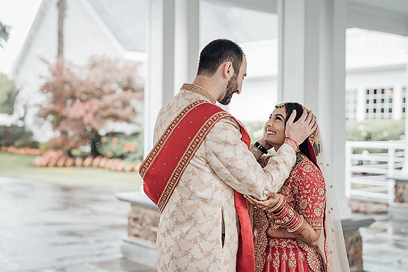 Couple portrait of an Indian bride and groom embracing under a covered porch with white columns, her red lehenga and maang tikka visible
