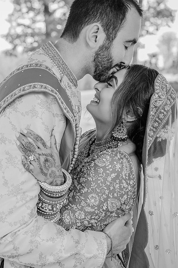Wedding couple portrait with groom kissing bride’s forehead as they embrace in embroidered sherwani and lehenga, trees softly blurred behind them