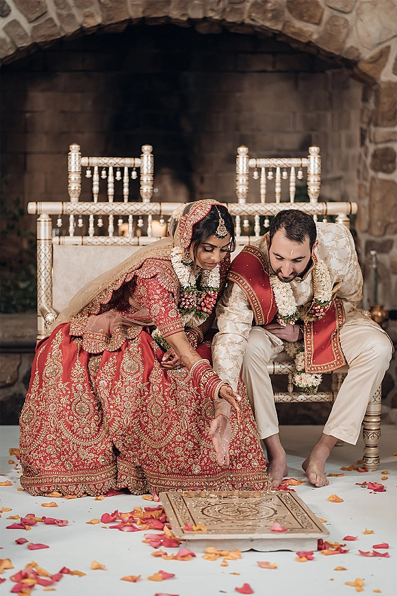 Hindu wedding ritual as Indian bride and groom sit on ornate chairs in red attire, wearing garlands and jewelry by a stone fireplace