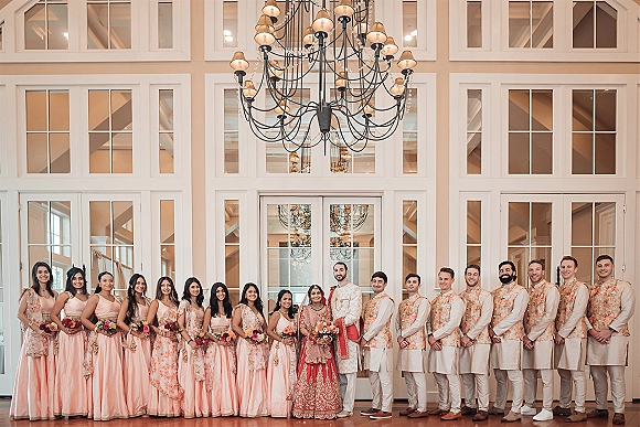 Wedding party portrait of bride and groom with wedding party lined up indoors before tall windows and double doors, under a chandelier