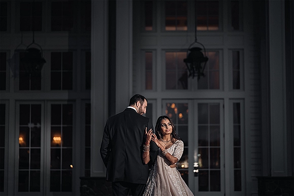 Couple portrait of bride holding groom’s arm in black-tie attire, her henna and veil highlighted by lantern-lit windows at night