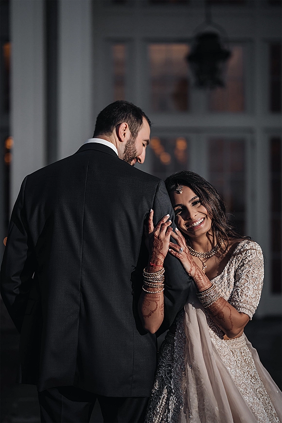 Couple portrait of a South Asian bride holding the groom’s arm, showing mehndi and bridal jewelry, by large windows in evening light