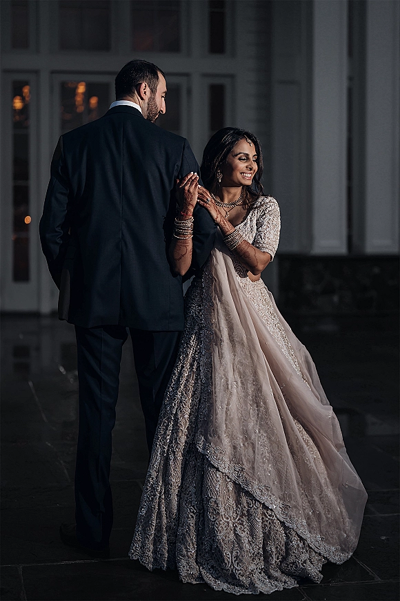 Couple portrait of a south asian wedding couple, bride holding groom’s arm showing mehndi, in lehenga by window panes and stone wall