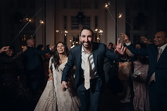 Wedding sparkler exit as bride in embellished lehenga and groom in suit run through guests holding sparklers at an indoor doorway at night
