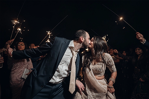 Wedding kiss during a sparkler send off as guests form a glowing tunnel at night, bride in lehenga with dupatta and jewelry