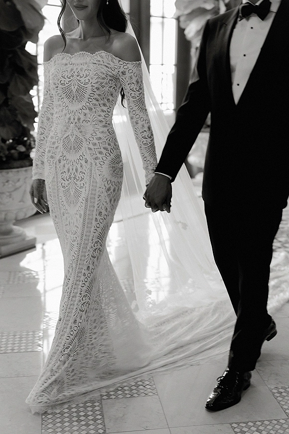 Couple portrait of bride and groom holding hands, bride in lace wedding dress with veil, groom in black tuxedo by tall windows indoors