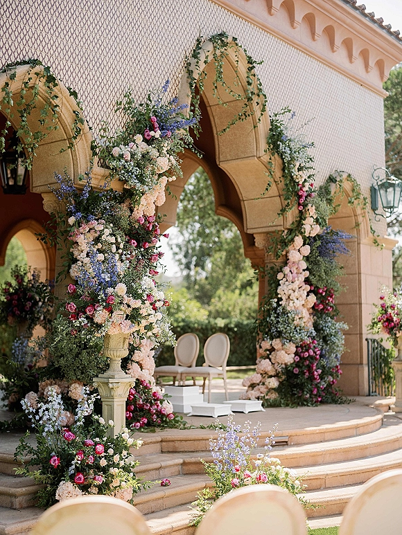Ceremony altar decor featuring a floral arch wedding of roses, hydrangeas and delphinium with greenery garland on stone steps by a colonnade