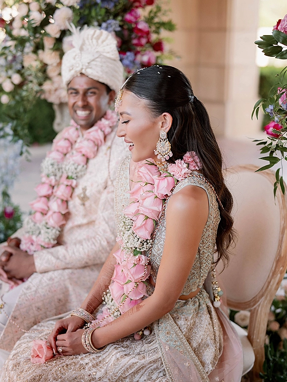 Wedding couple portrait of a South Asian bride and groom seated on a floral stage, wearing pink rose garlands and traditional attire