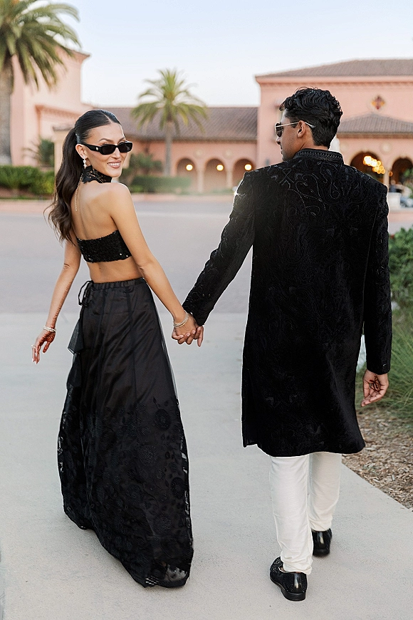 Couple portrait of bride and groom holding hands in black wedding outfits, walking away as she looks over shoulder in a palm-lined courtyard walkway