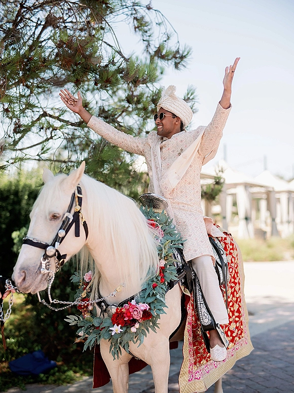 Baraat groom entrance with groom on white horse, wearing cream sherwani, safa turban, and sunglasses under leafy trees outdoors