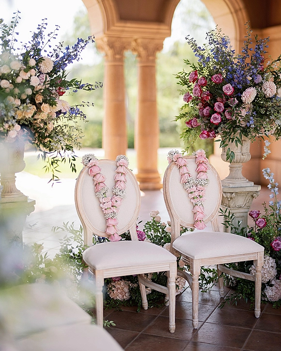 Ceremony chairs decor with white oval-back chairs draped in pink rose garlands and baby's breath beneath stone arches and greenery backdrop