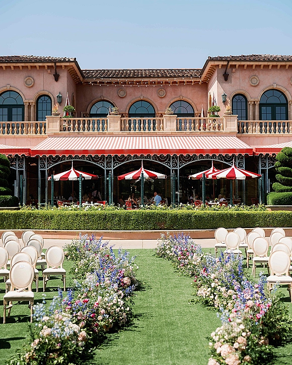 Ceremony aisle decor with outdoor ceremony aisle flowers in low meadow-style clusters along grass, facing terrace with arched windows and striped umbrellas