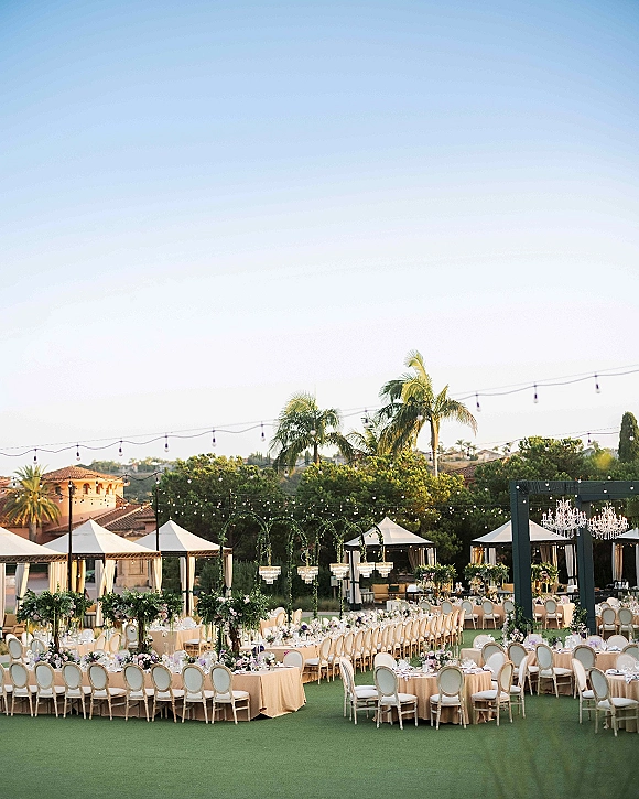 Outdoor reception setup with round banquet tables in cream linens, pastel floral centerpieces, white oval-back chairs, chandeliers and string lights on a lawn with palms