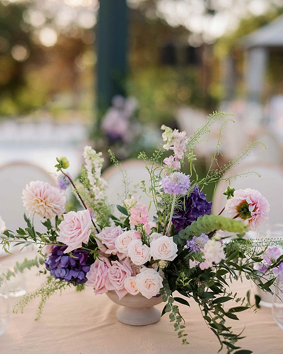 Wedding centerpiece featuring pastel roses, dahlias, and hydrangea in a compote vase on a tablecloth at a sunlit garden reception