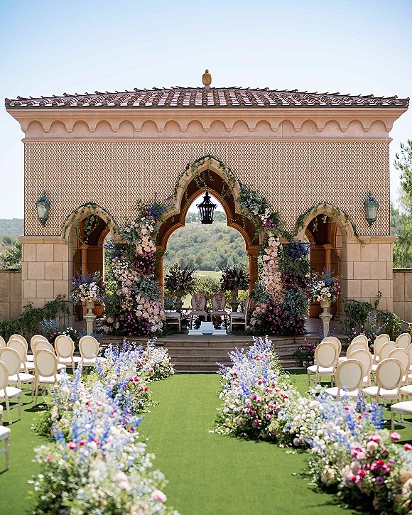 Ceremony setup with an outdoor wedding ceremony floral arch, aisle flowers and white chairs on a terrace by an arched facade and hills