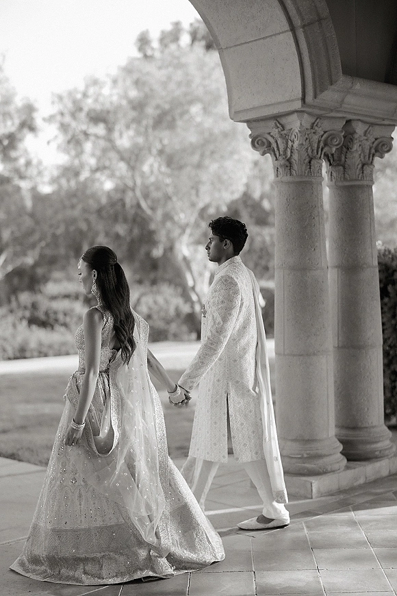Wedding couple portrait of a south asian wedding couple holding hands, walking under a stone archway with carved columns, bride in lehenga veil