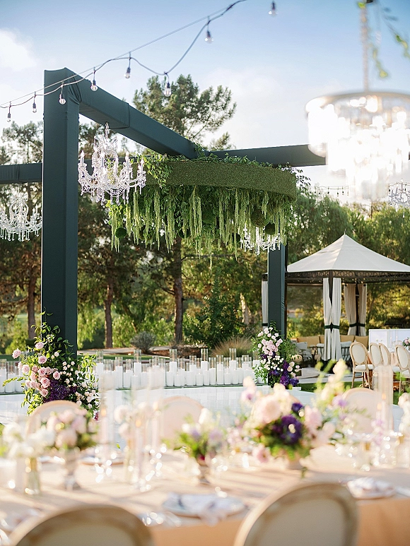 Outdoor reception decor with wedding chandelier decor, string lights and hanging greenery above white tables under a pergola with trees and blue sky