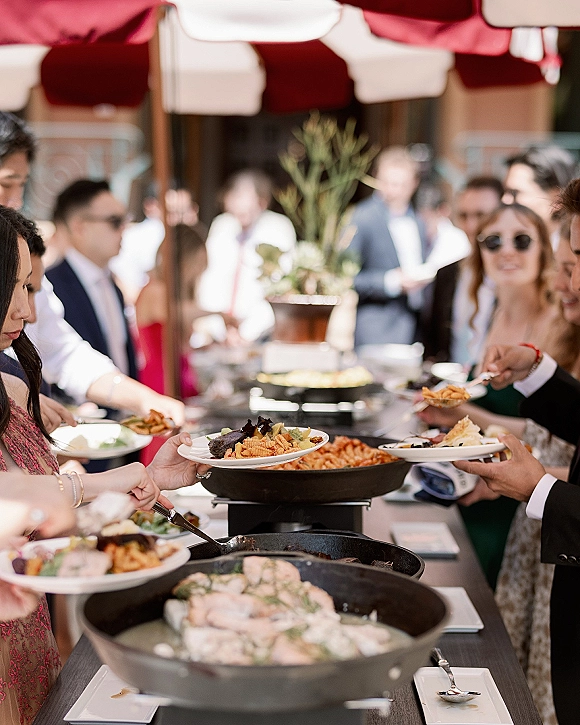 Wedding buffet line with wedding catering buffet chafing pans as guests in cocktail attire hold plates on an outdoor patio under red umbrellas