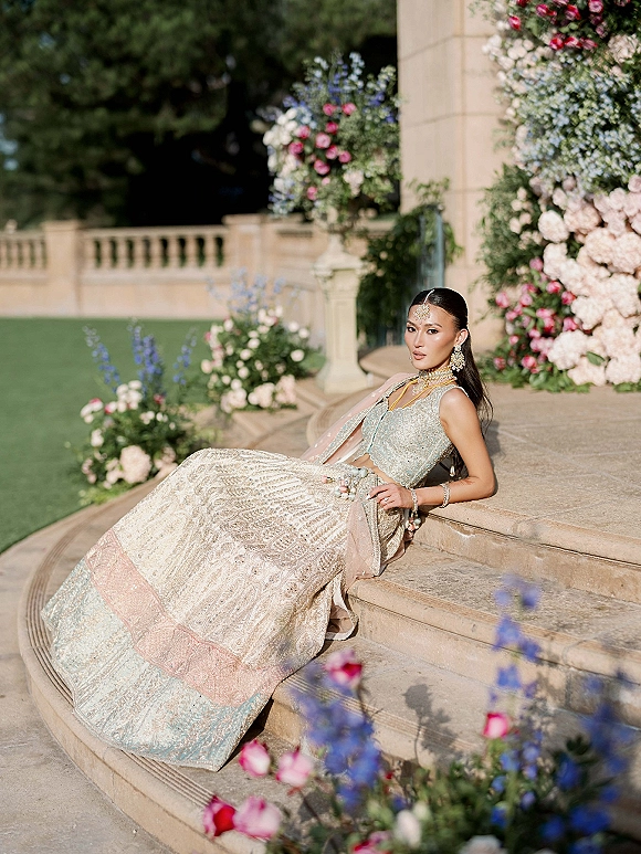 Bridal portrait of a South Asian bride in an embellished lehenga with gold jewelry, seated on stone steps amid garden greenery
