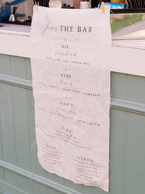 Bar menu sign with wedding bar menu calligraphy printed on linen fabric, clipped with wooden clothespins above a bar counter with cans and ice bucket