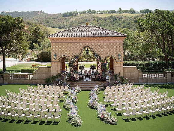 Ceremony setup with outdoor wedding ceremony floral arch and aisle florals, white chairs facing a stone pavilion under terracotta roof