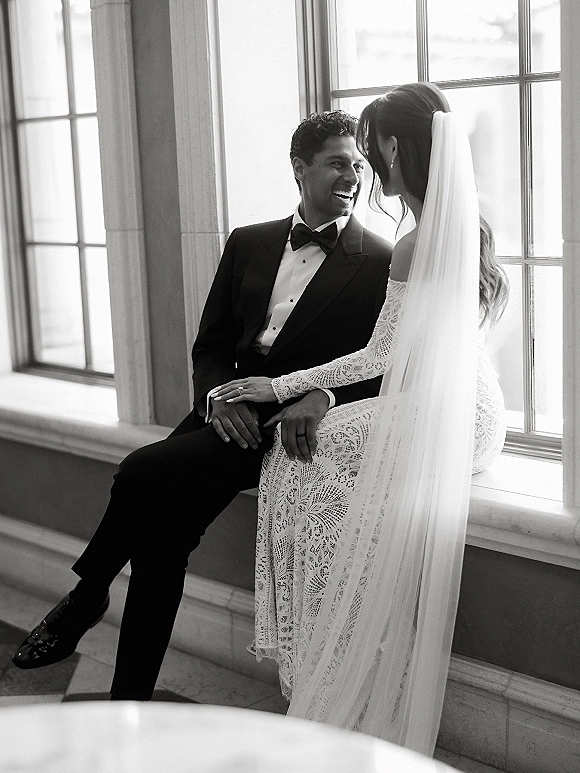 Couple portrait of bride and groom sitting by large windows in natural light, laughing and holding hands, veil and black tuxedo visible