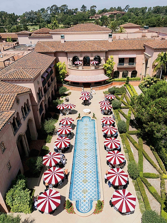 Outdoor reception setup with poolside wedding reception tables, striped umbrellas and lounge chairs in a Mediterranean villa courtyard under blue sky